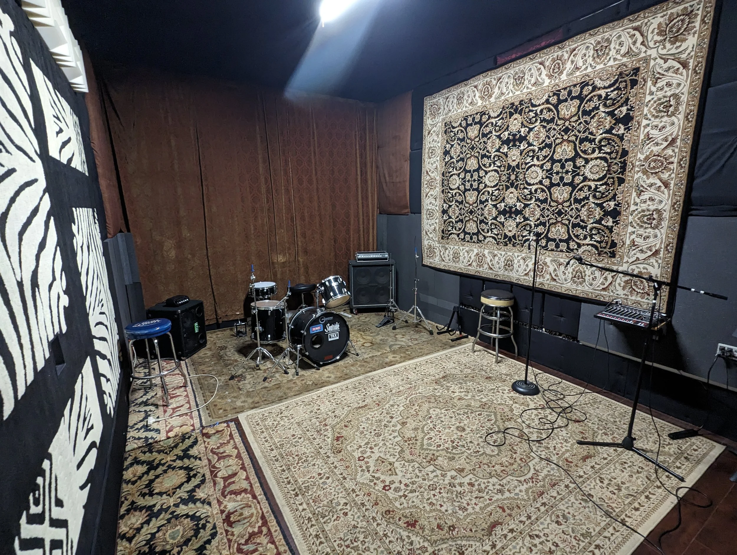 Small rehearsal studio with drums, backline, and PA. Tall ceilings with curtains, ornate rugs, and enough space for up to 5 people.