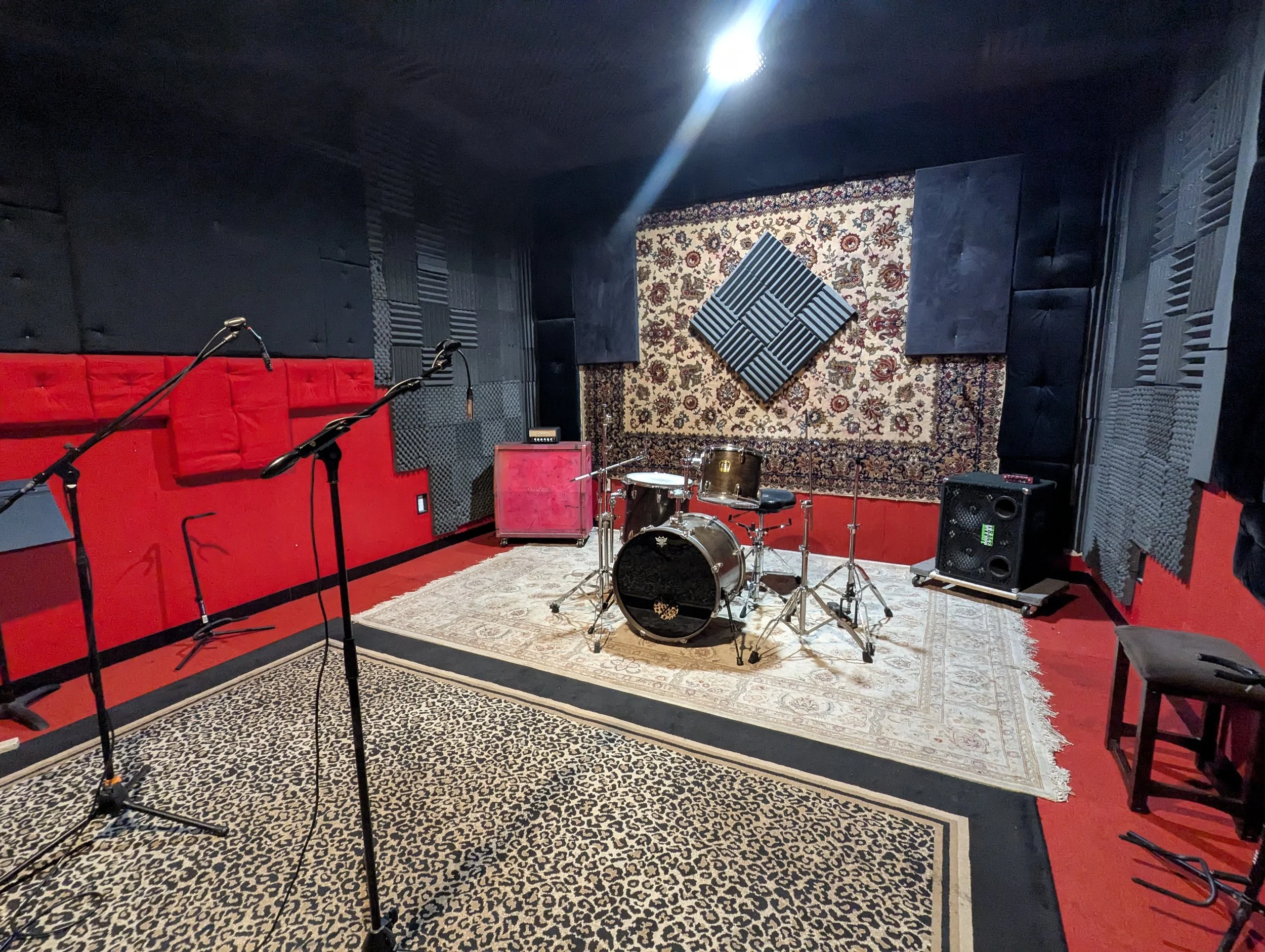 Medium rehearsal room with bright red walls, black auralex, and wild leopard print rug. Equipped with backline, drums, and PA.