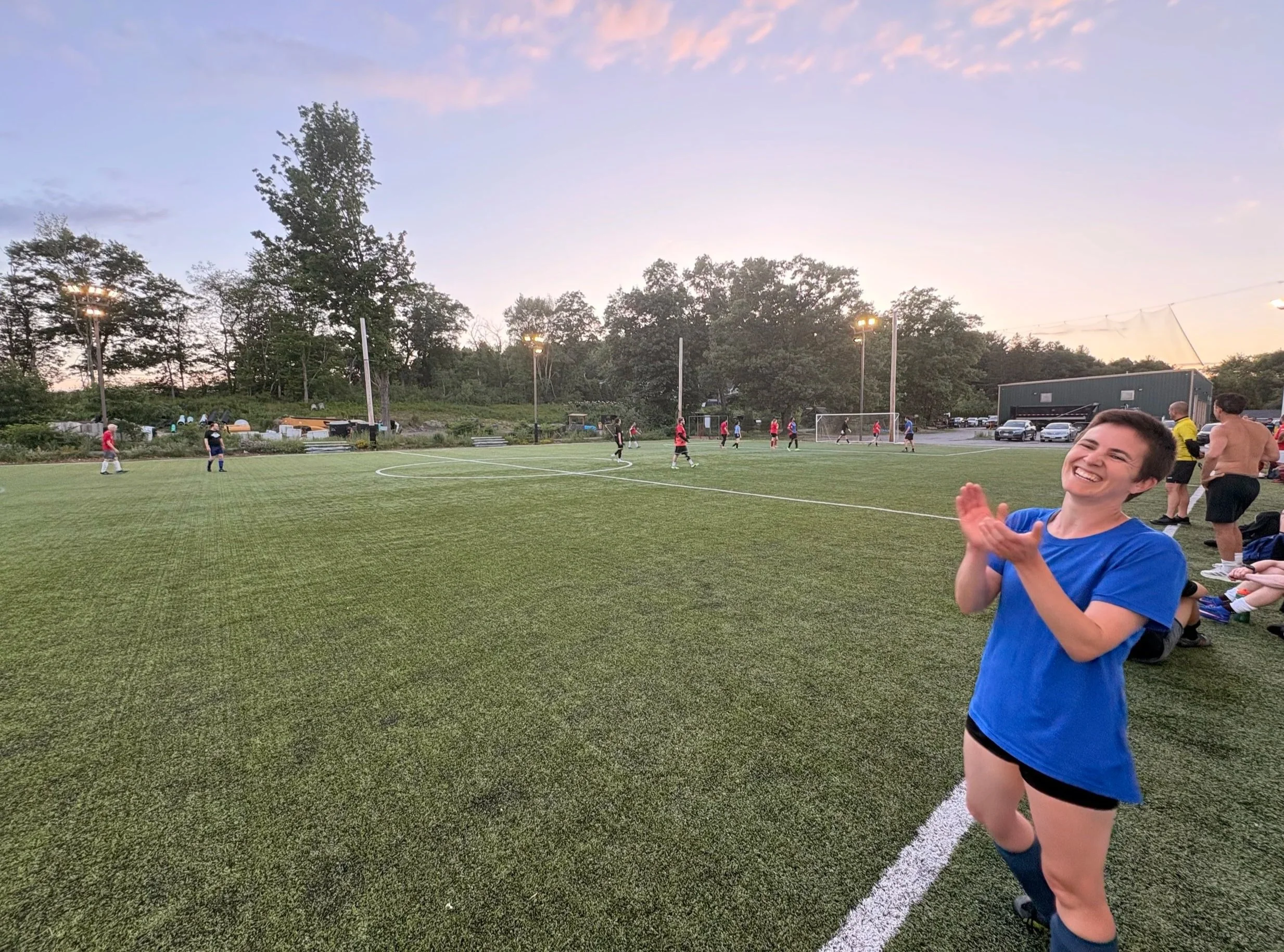 woman in blue T-shirt and soccer shoes clapping at the sidelines of a soccer field