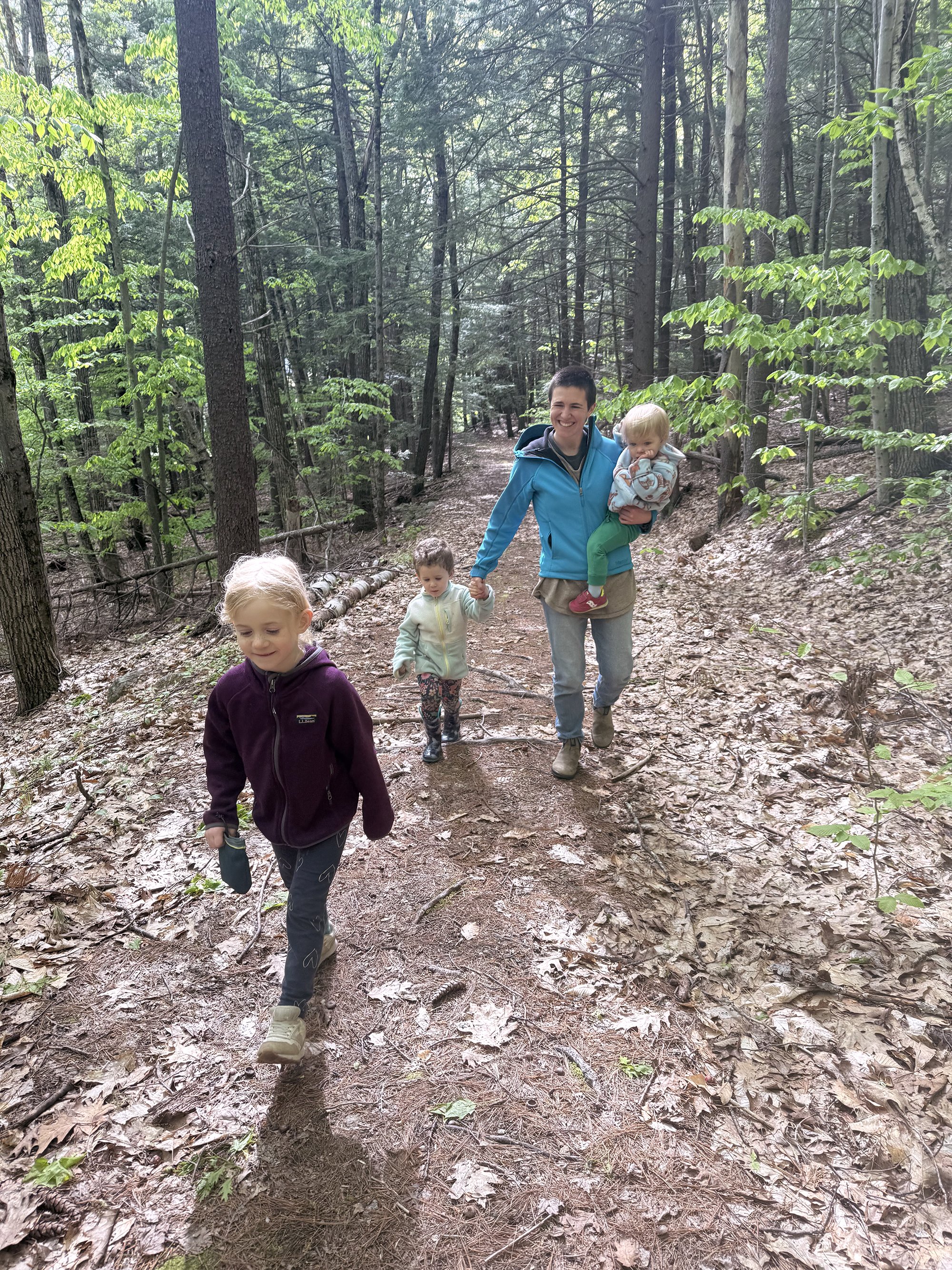 a woman in jeans walking through a wooded trail with 3 children, the smallest one on her hip