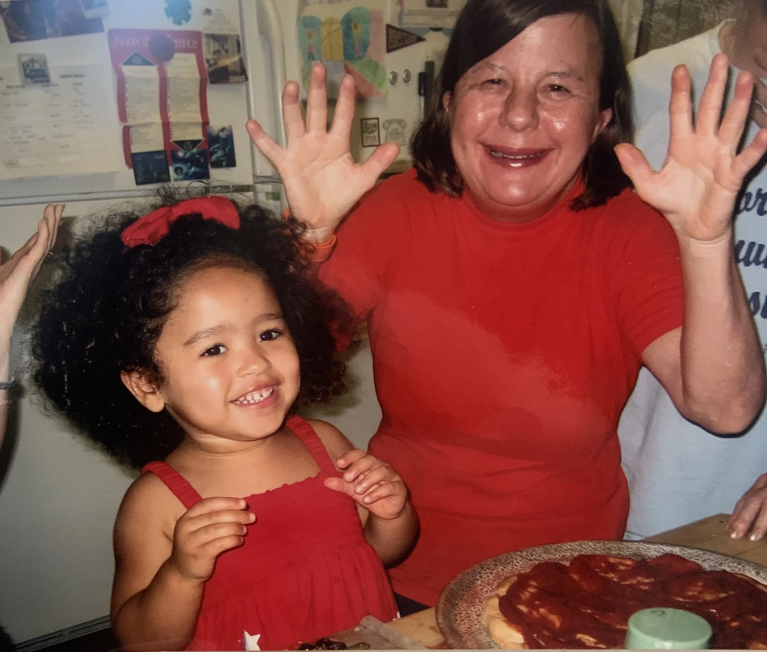 a young girl with curly hair and a red dress smiles next to an adult woman in a red t-shirt smiling widely.