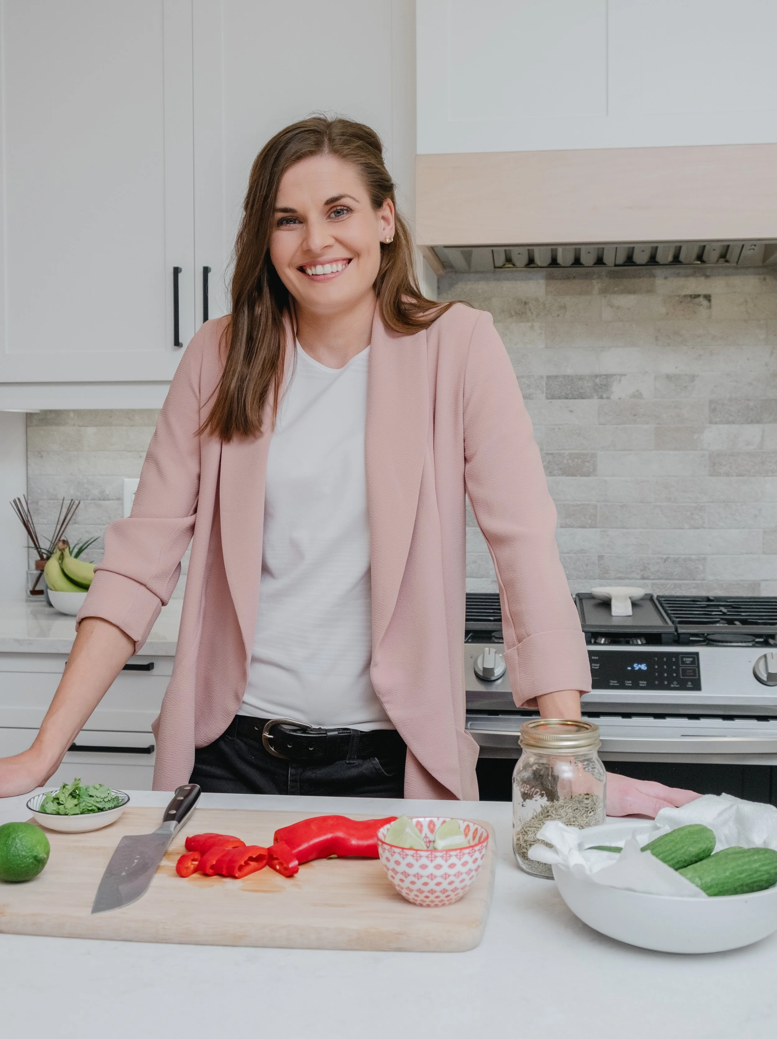A woman standing in a kitchen, smiling, with a cutting board, knife, and vegetables like red bell pepper, lime, and cucumbers on the counter.