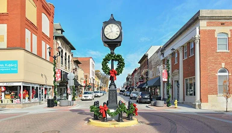 Historic downtown street with a decorative clock monument, brick buildings, storefronts, and parked vehicles.