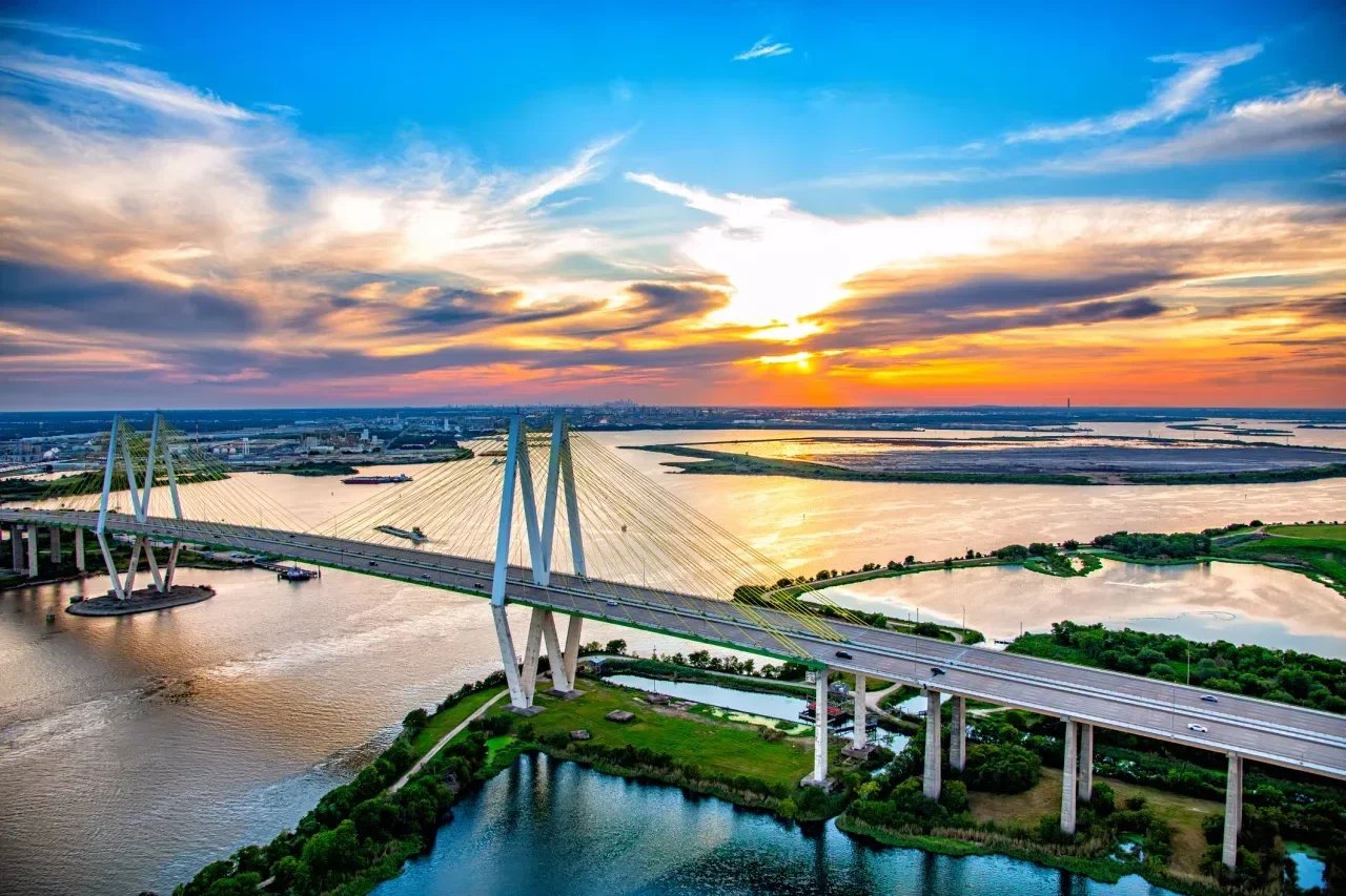 Aerial view of a cable-stayed bridge over a river at sunset, with colorful clouds, wetlands, and skyline in distance.