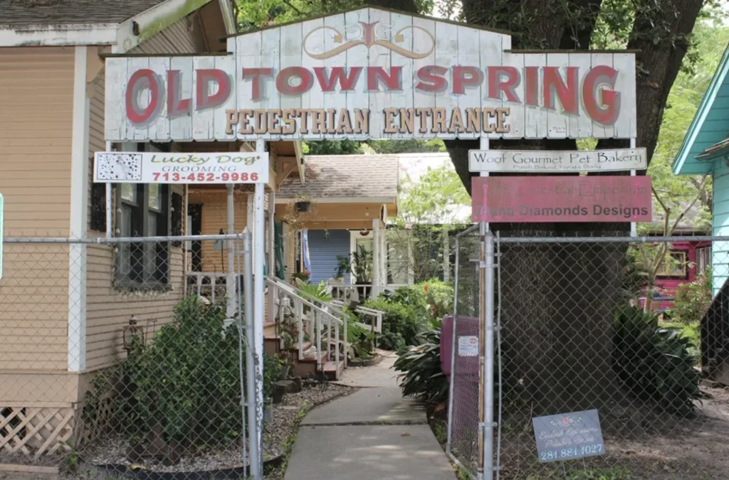 Old Town Spring pedestrian entrance sign above a walkway lined with small shops, fencing, trees, and greenery.