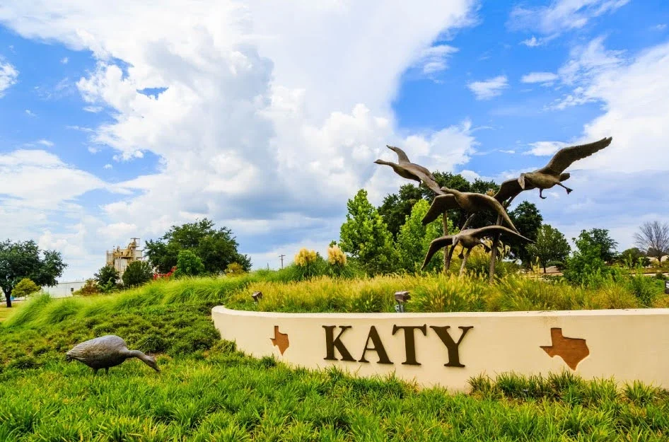 Katy Texas city sign with geese statues, landscaped greenery, and dramatic cloudy sky in park setting.