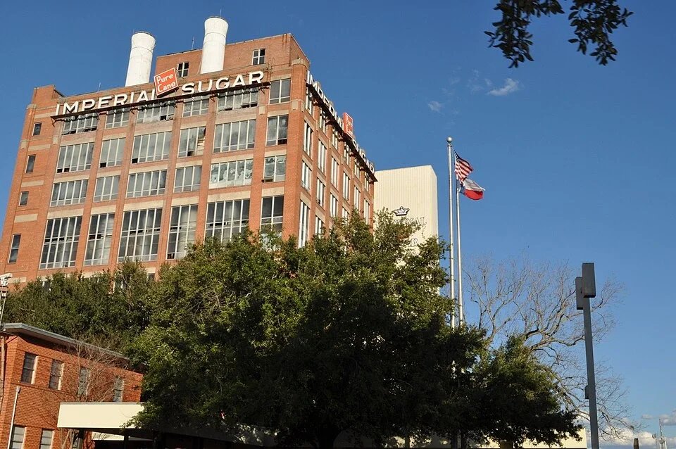 Historic Imperial Sugar factory building with brick facade, industrial features, and flags beneath a clear sky.