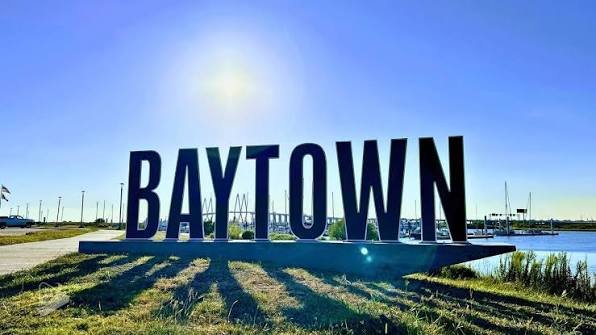 Large BAYTOWN sign beside a waterfront marina under a clear blue sky, with sunlight casting long shadows on grass.