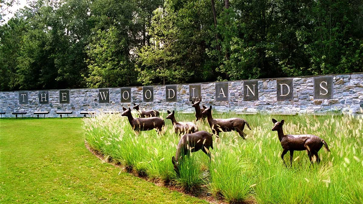 The Woodlands Texas entrance sign with deer statues, stone wall, landscaped grass, and trees in background.