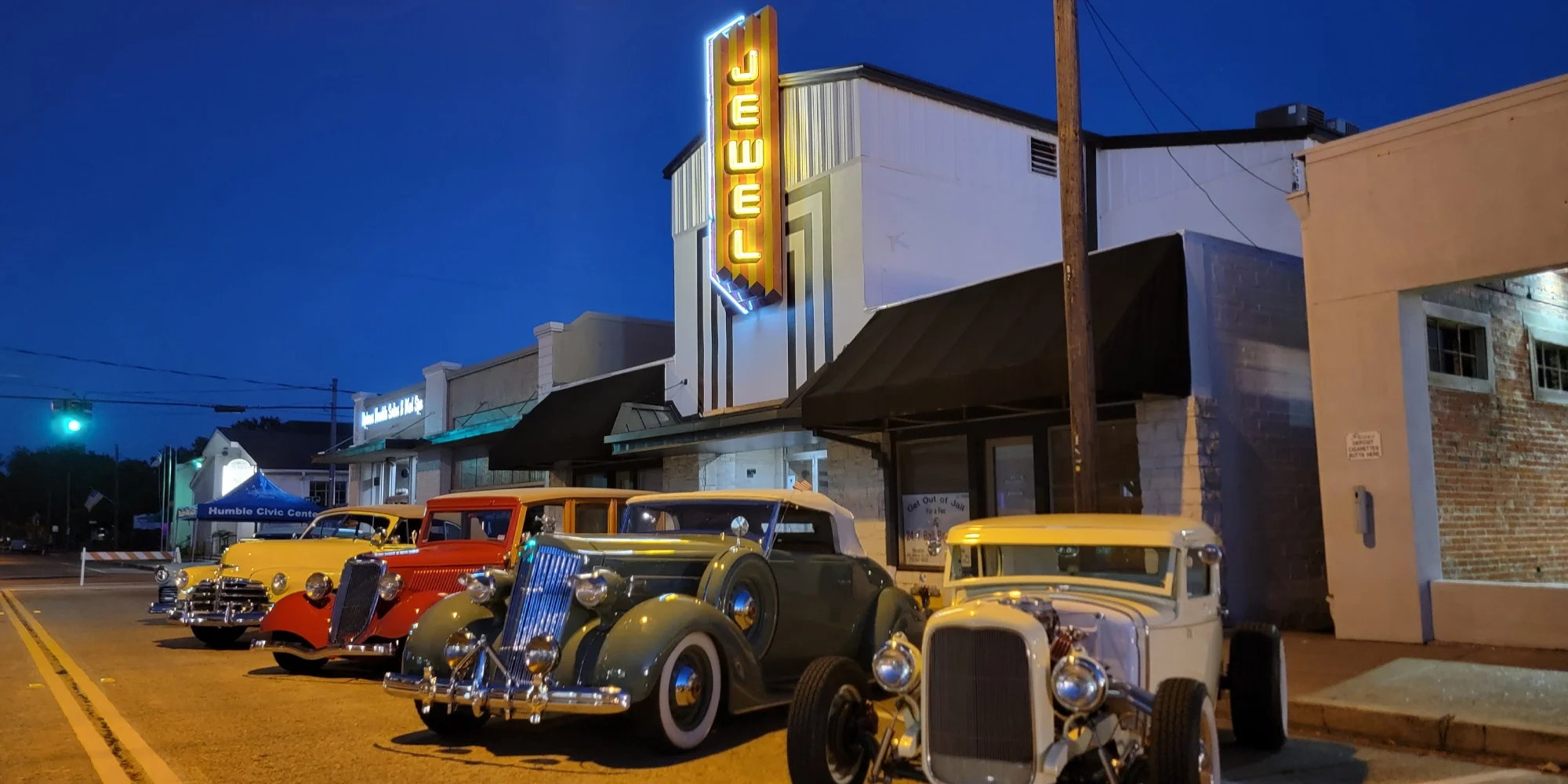 Vintage classic cars parked on a street at night in front of the Jewel Theater with a bright neon sign and storefronts.