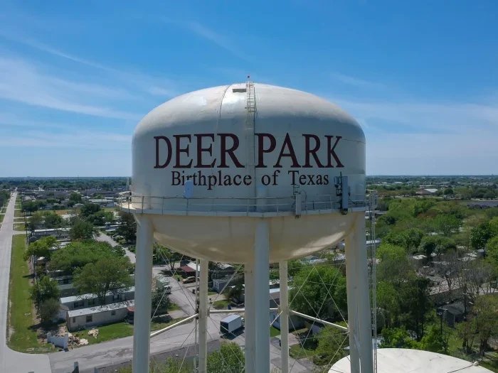 Water tower labeled Deer Park Birthplace of Texas rising above a residential area under a clear blue sky.