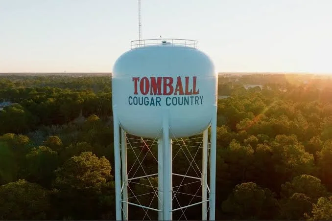 Water tower labeled Tomball Cougar Country rising above a wooded landscape at sunset, viewed from an aerial angle.