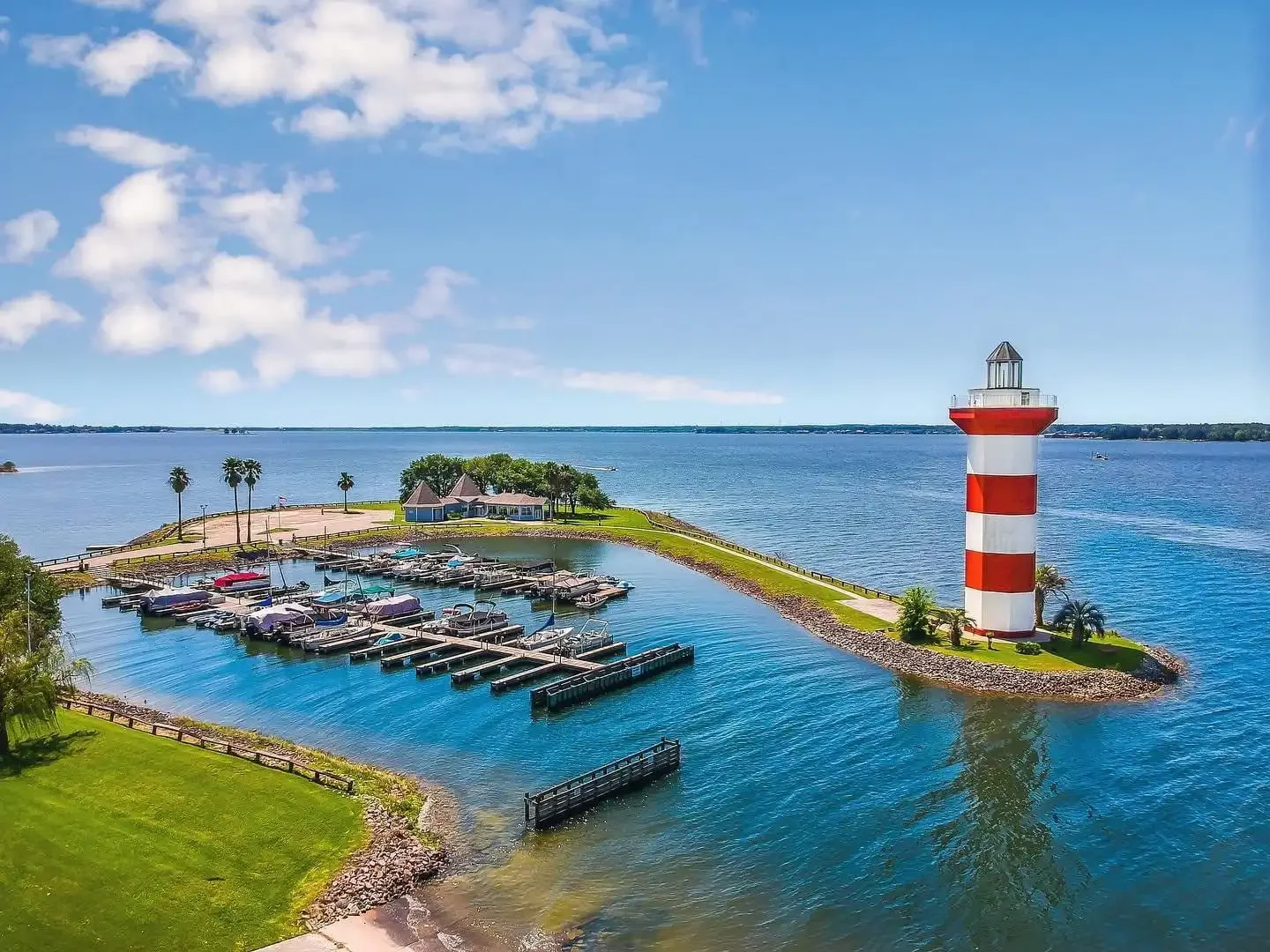 Aerial view of a marina and striped lighthouse on a lakeside peninsula, with docks, boats, and calm blue water.