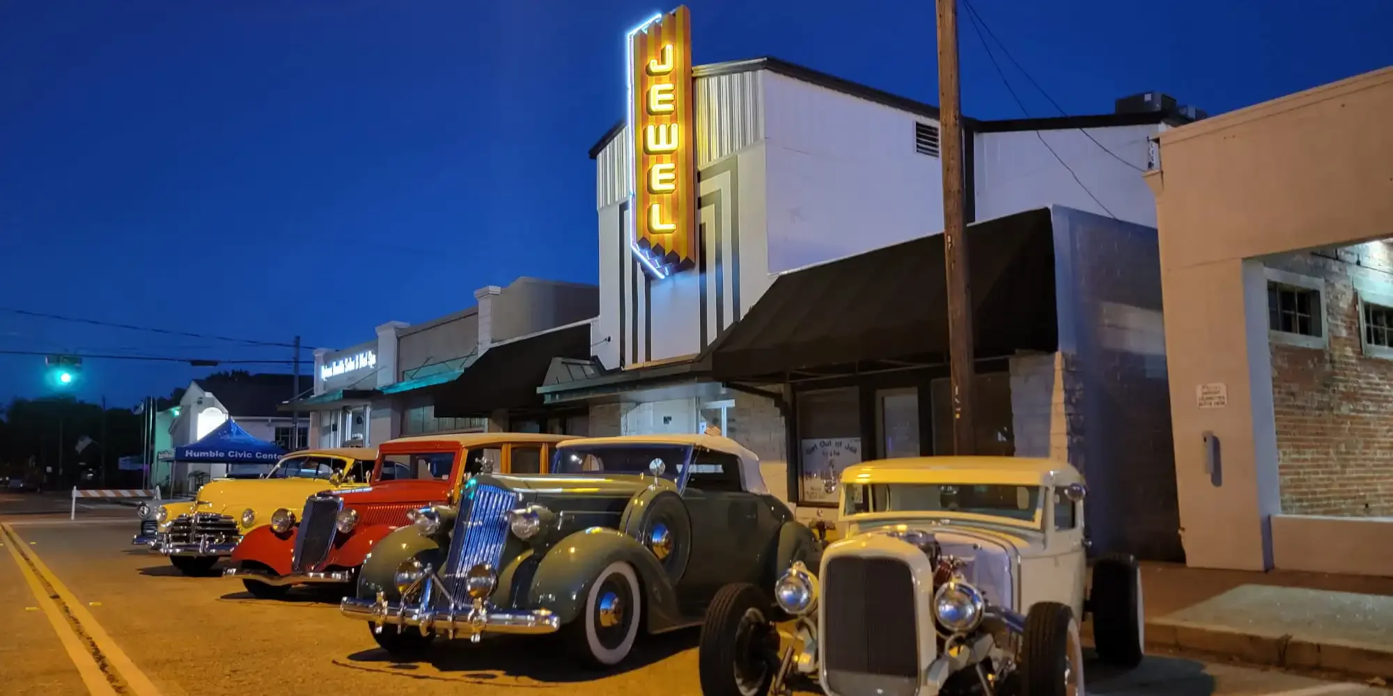Vintage cars parked outside the historic Jewel Theater at night, with a glowing marquee in a downtown street scene.