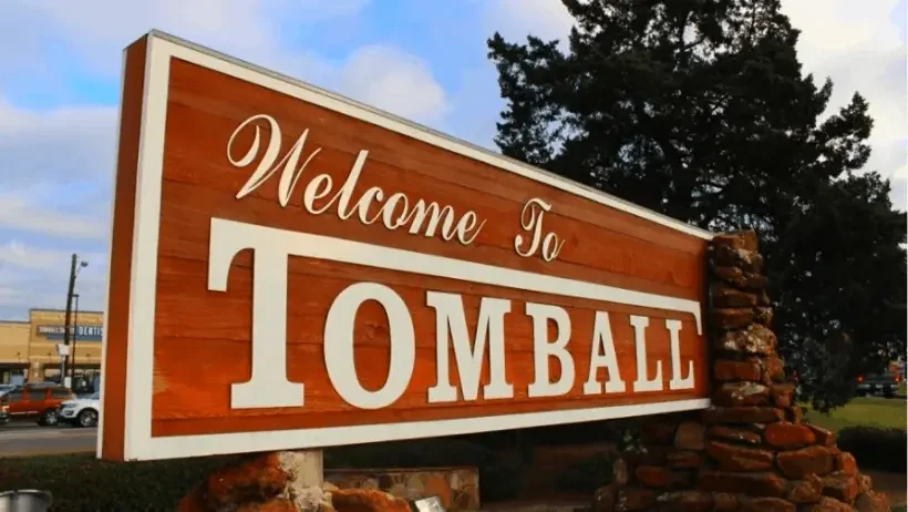 Large Welcome to Tomball sign with wood panel design and stone accents near a roadside entrance under cloudy skies.