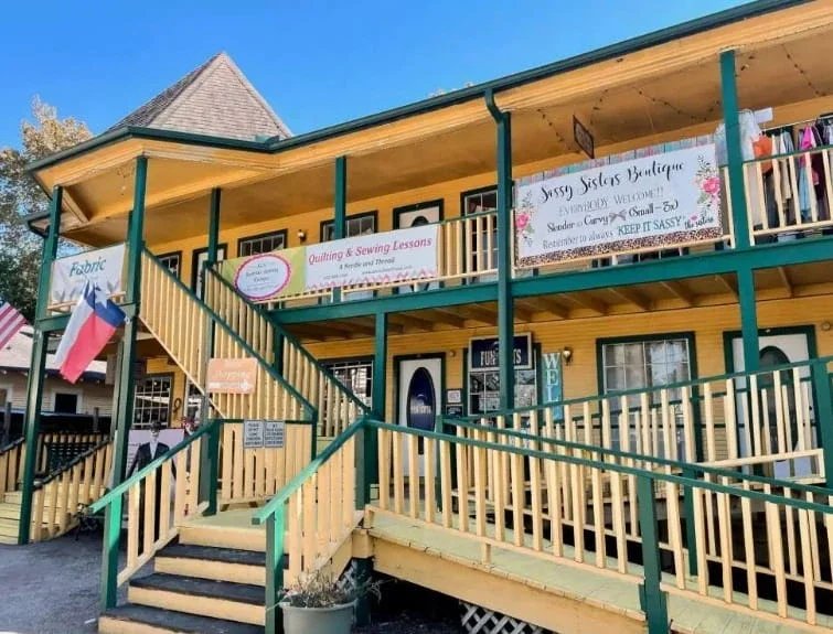 Two-story historic retail building with exterior stairs, balconies, shop signs, and a Texas flag under clear skies.