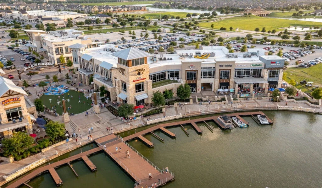 Aerial view of a lakeside retail and dining center with boardwalk docks, storefronts, parking lots, and green space.