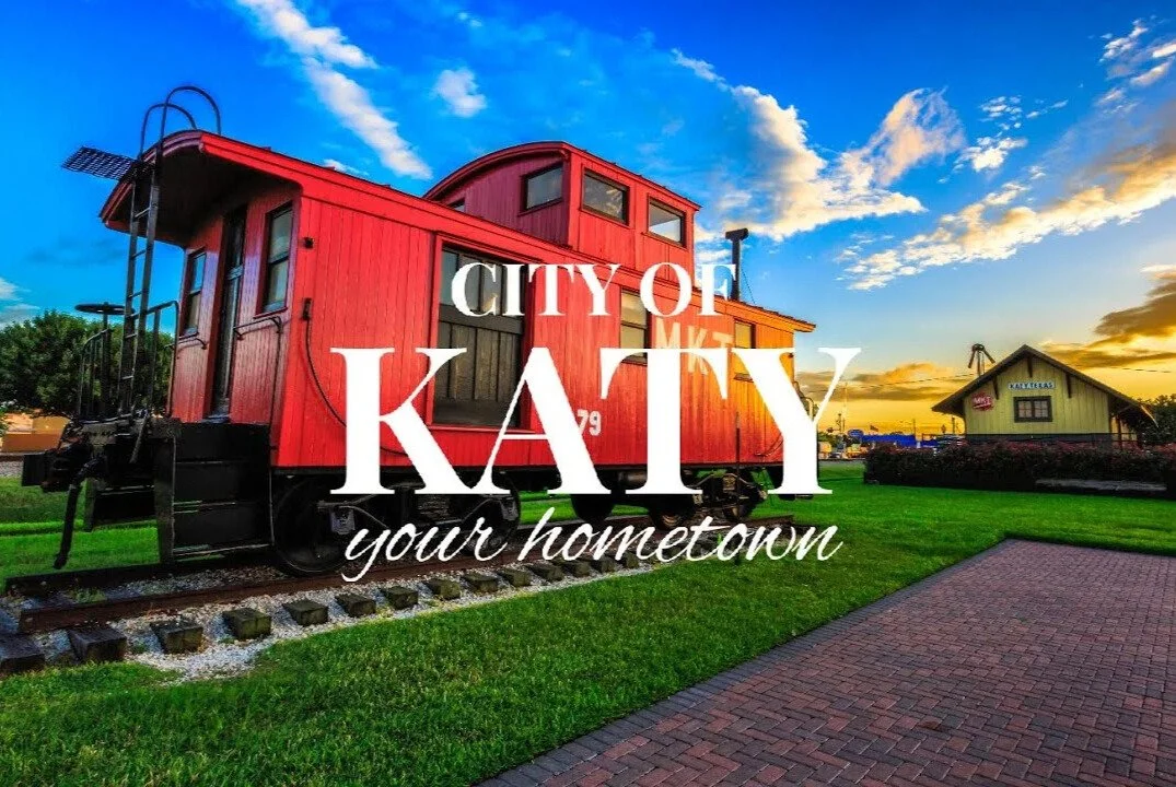 City of Katy Texas red caboose train display in park at sunset with brick path and historic depot building in background.