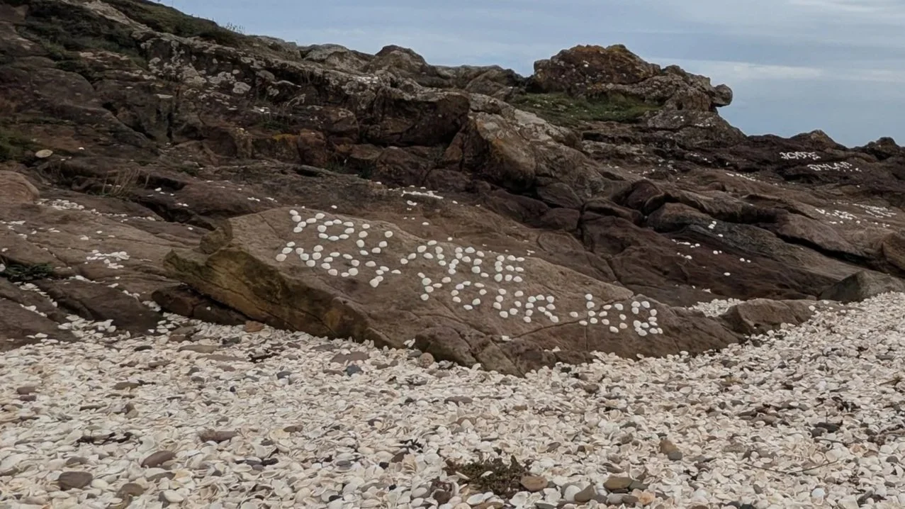 A rocky beach with lots of clam shells. Some of the shells are arranged on one of the rocks to spell the message 'you are not your job'.