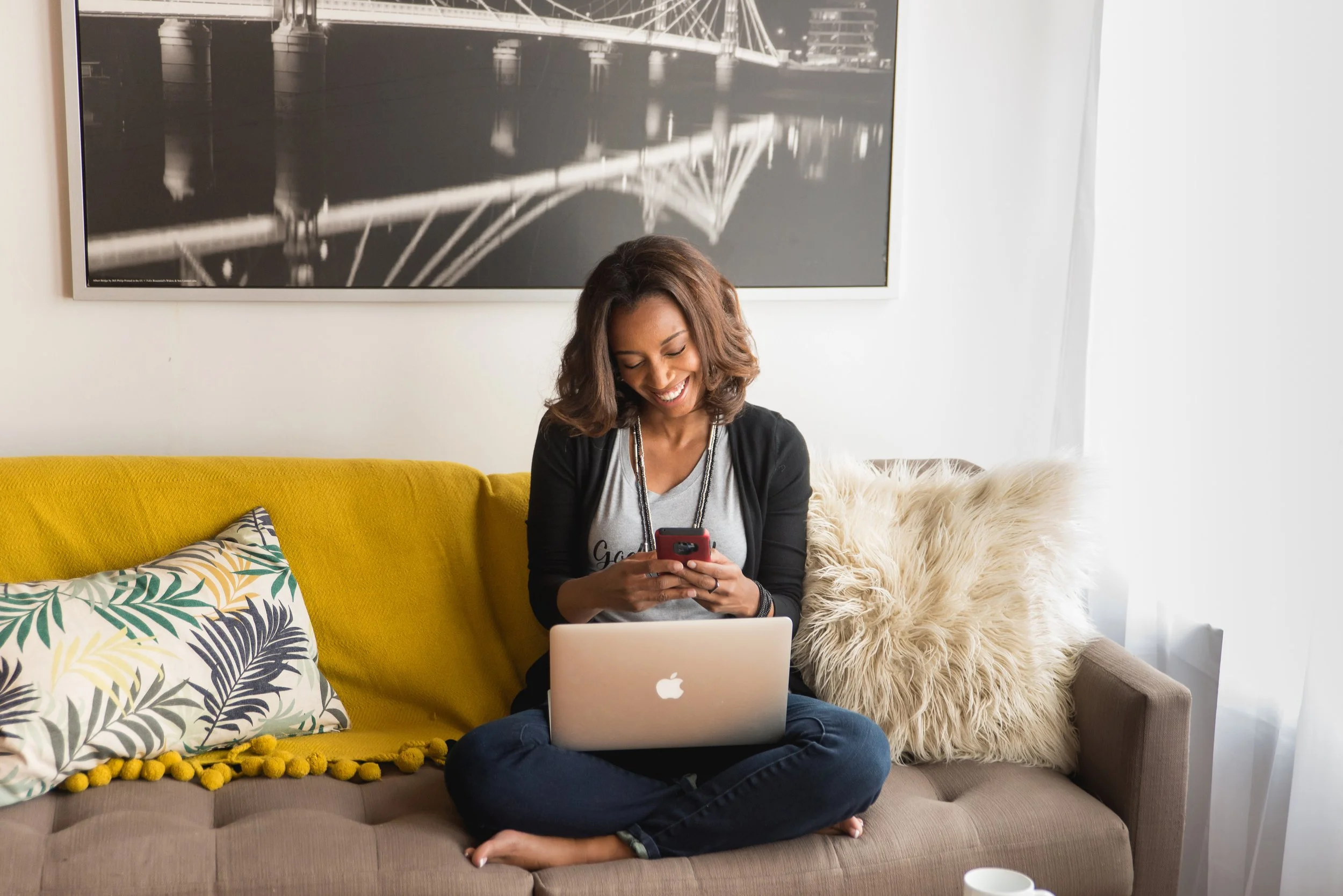 A woman sitting cross-legged on a sofa, working on a laptop and using her smartphone, in a bright living room with white curtains and a large black and white cityscape photograph on the wall.