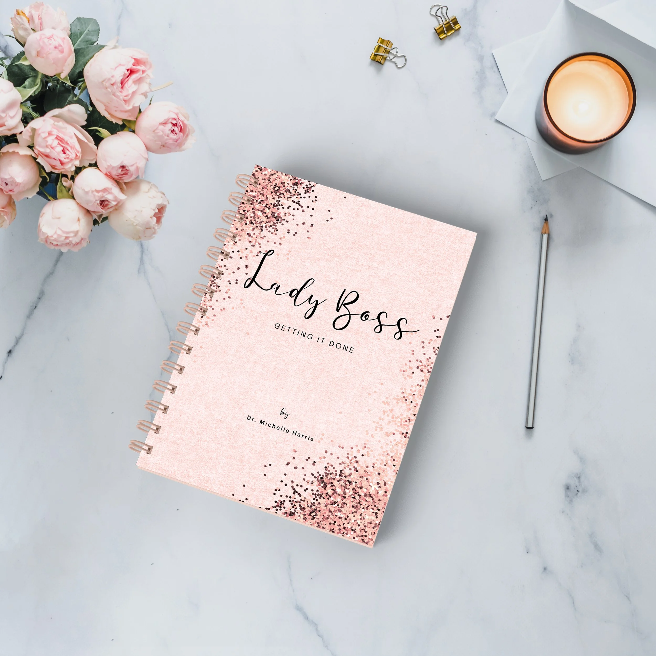A pink notebook titled 'Lady Boss Getting It Done' is on a marble surface. Beside it is a bouquet of pink peonies, a lit candle, a silver pencil, and a white envelope with gold binder clips.
