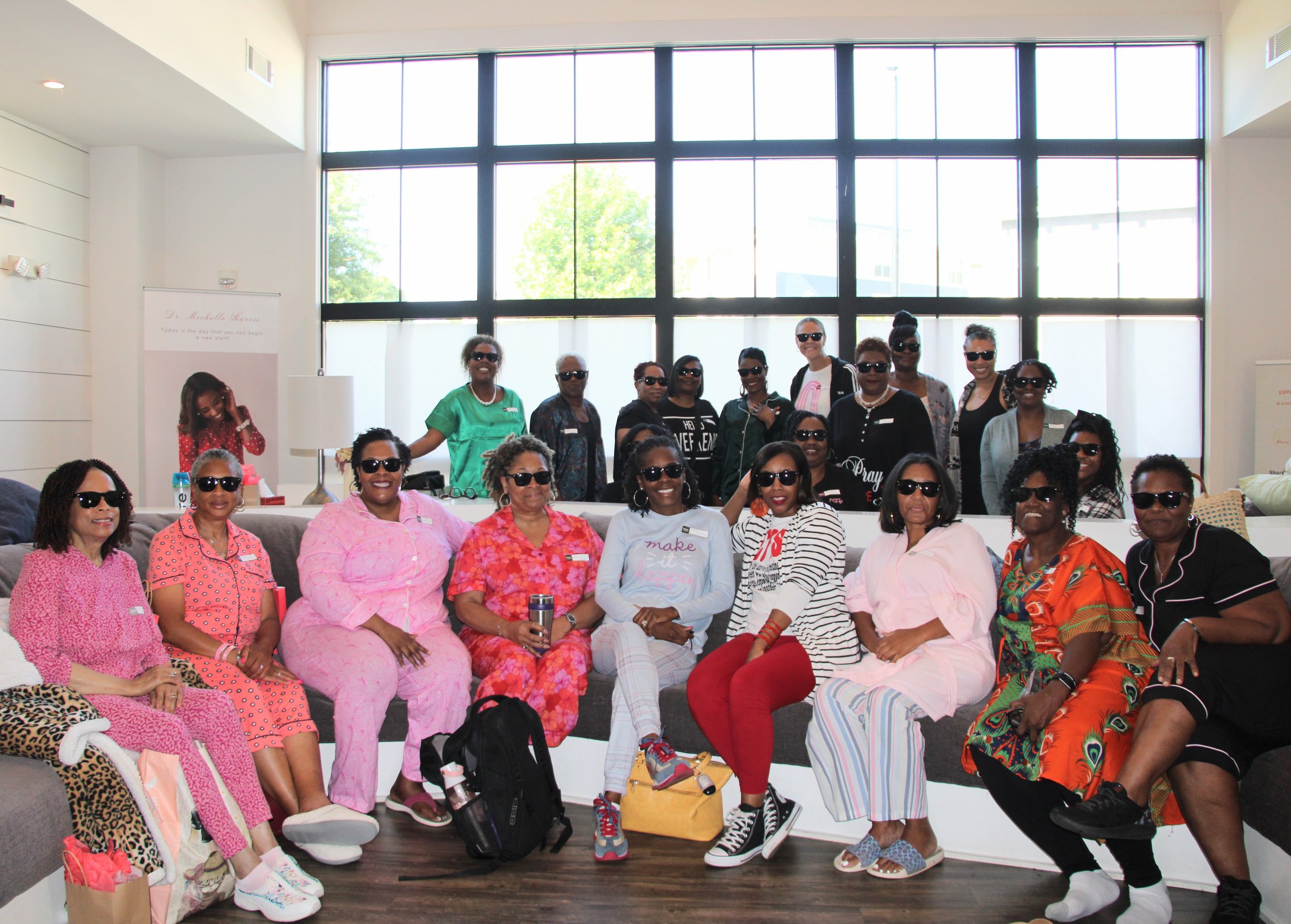 Group of women wearing sunglasses, seated and standing in a brightly lit indoor space with large windows.