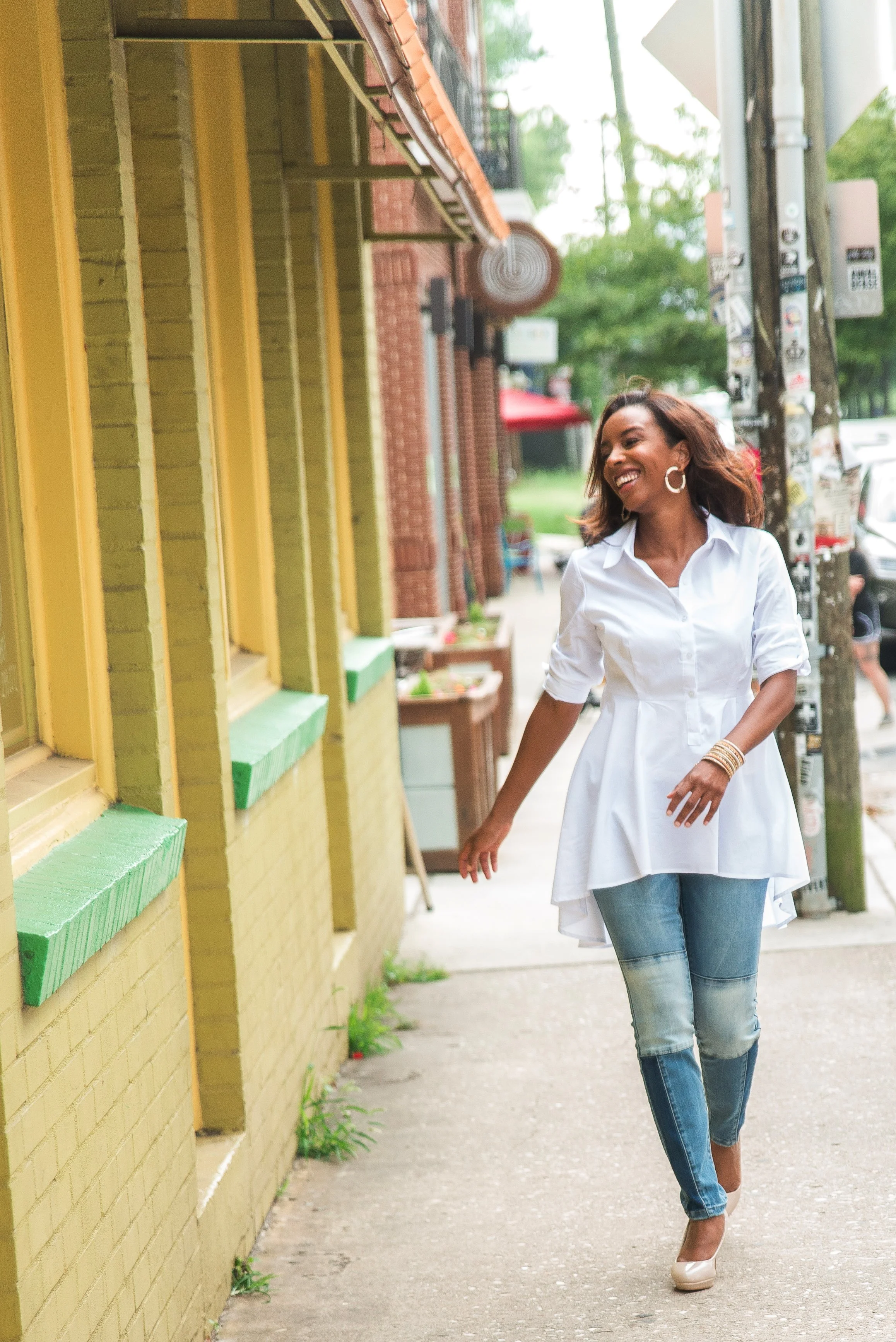 A woman with shoulder-length hair wearing a white dress, jeans, and high heels walking happily along a sidewalk in an urban area.