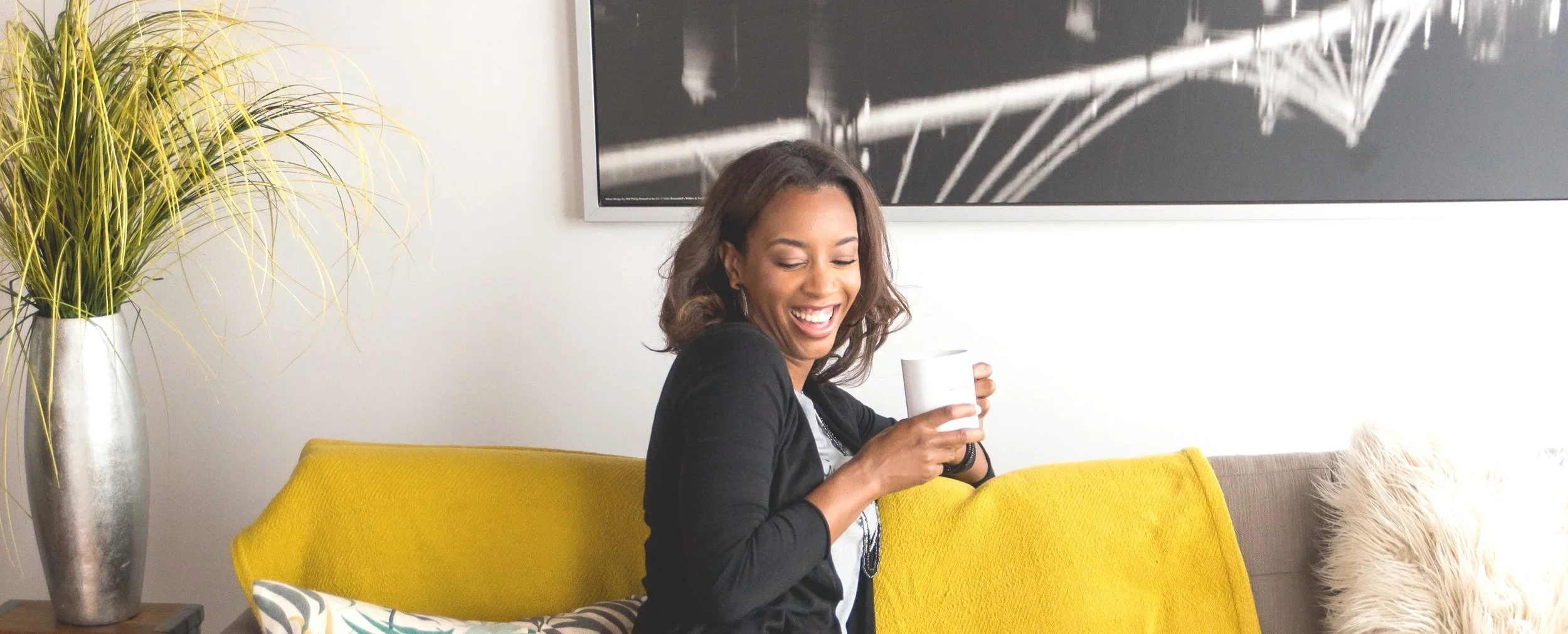 A woman sitting on a yellow sofa holding a white mug, smiling and looking down, in a modern living room with a large black and white wall art, a tall silver vase with green plants, and a fluffy throw pillow.