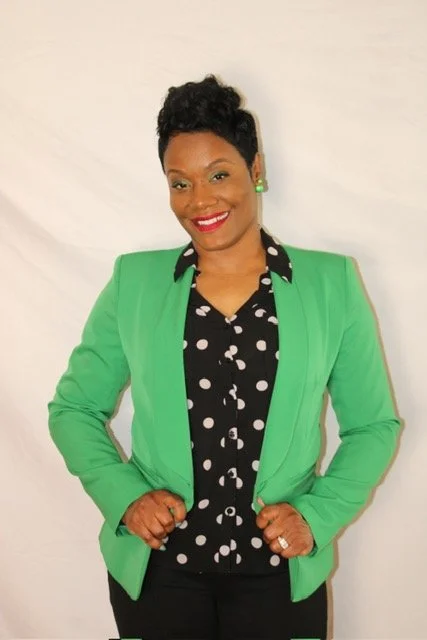 A woman with short curly black hair, wearing a bright green blazer over a black blouse with white polka dots, smiling, standing against a plain white background.