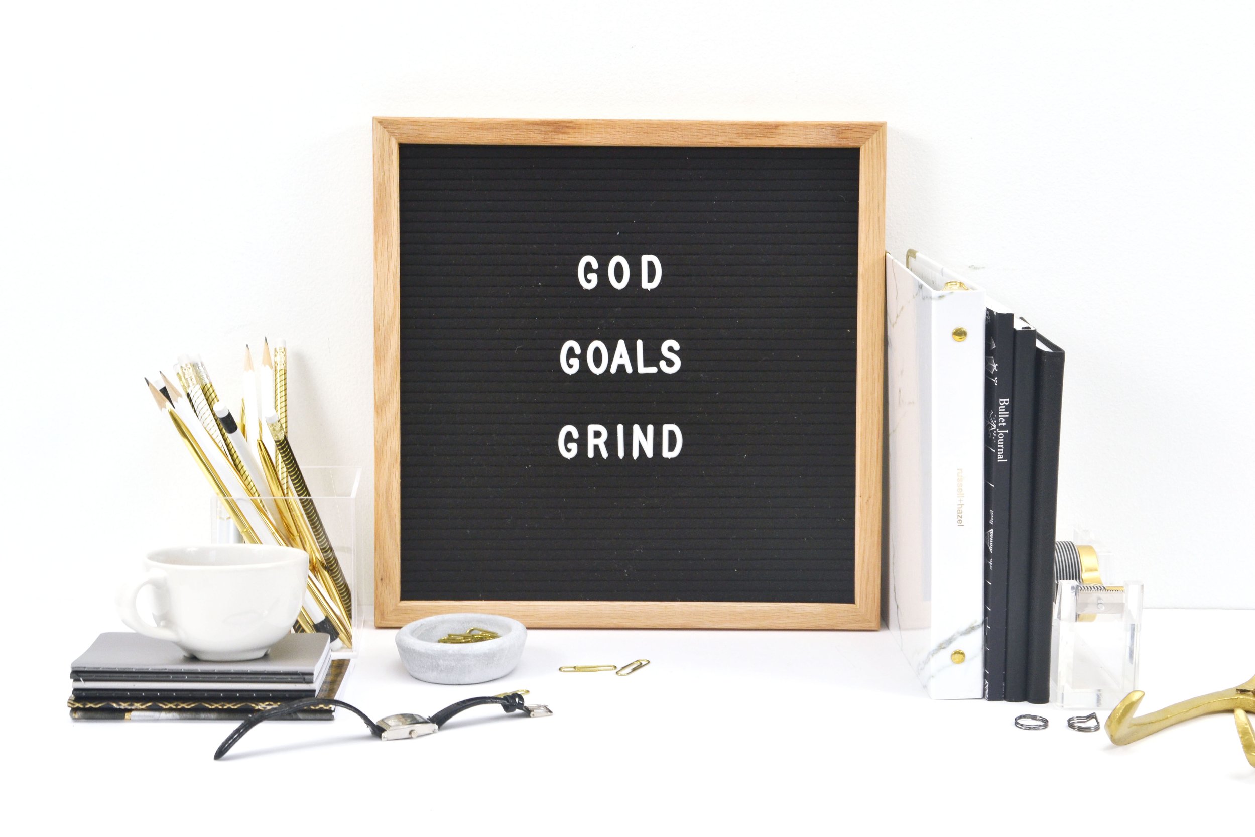 Desk setup with a blackboard that says 'GOD GOALS GRIND', notebooks, gold pens, a white mug, a watch, paper clips, and various office supplies.