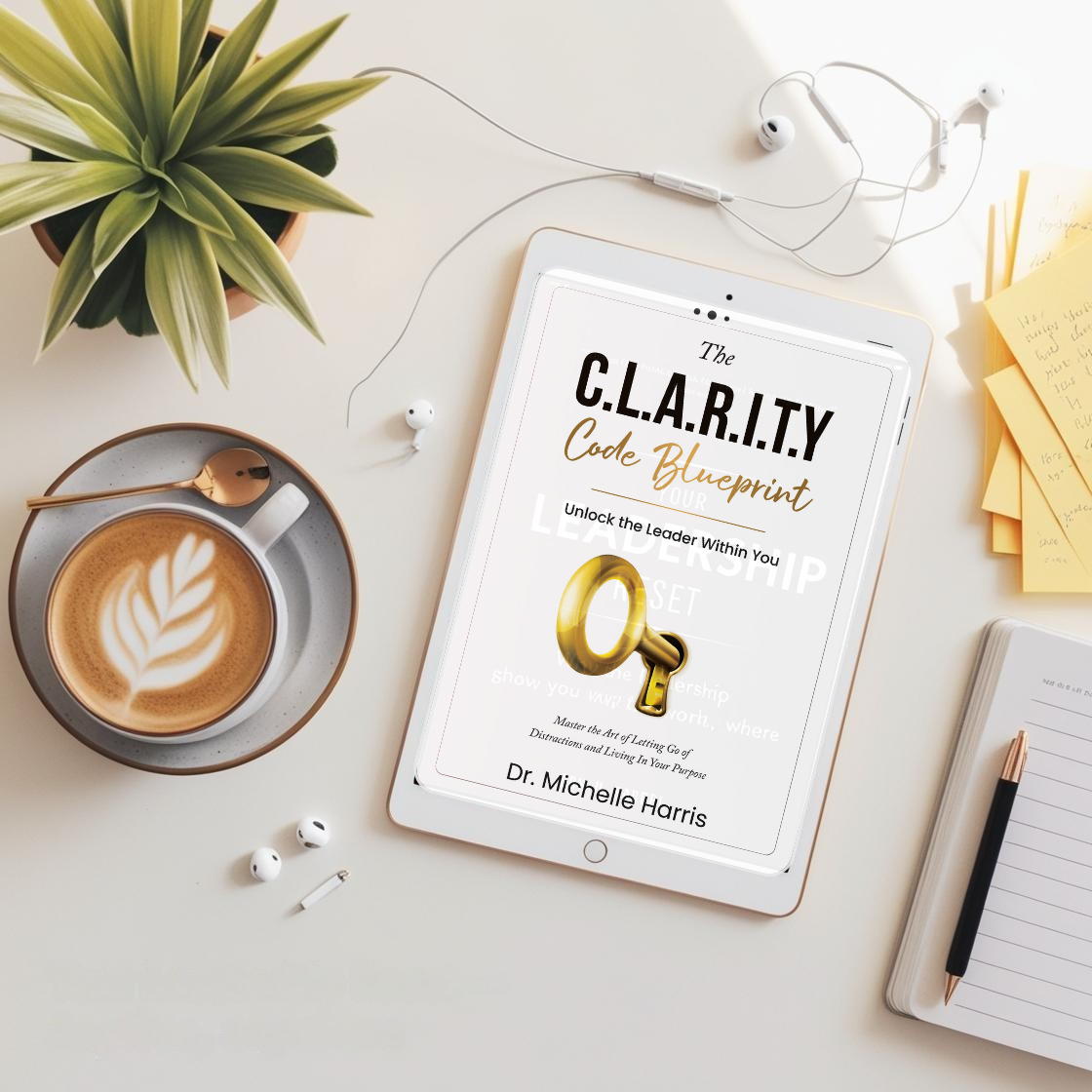 Flat lay of a workspace with a tablet displaying a book titled "The C.L.A.R.I.T.Y Code Blueprint" by Dr. Michelle Harris, a cup of latte with latte art, a gold key, white earbuds, sticky notes with handwritten notes, a notebook with a pen, and a potted plant.