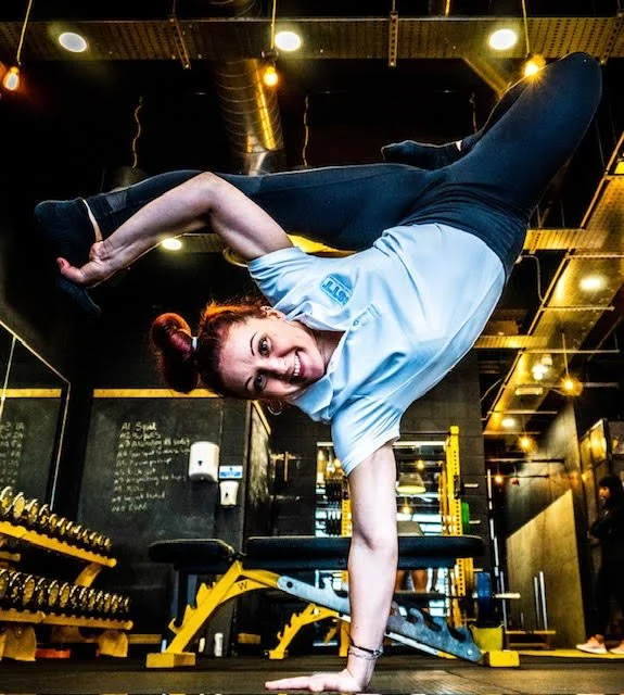 Woman with red hair performing a one-handed handstand in a gym at BSTT, demonstrating strength, flexibility, and athletic performance