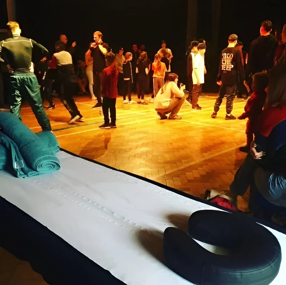People dancing and socializing in a dimly lit room with wooden floor, viewed from a massage table at BSTT, illustrating Hip Hop Health and dance wellness programs