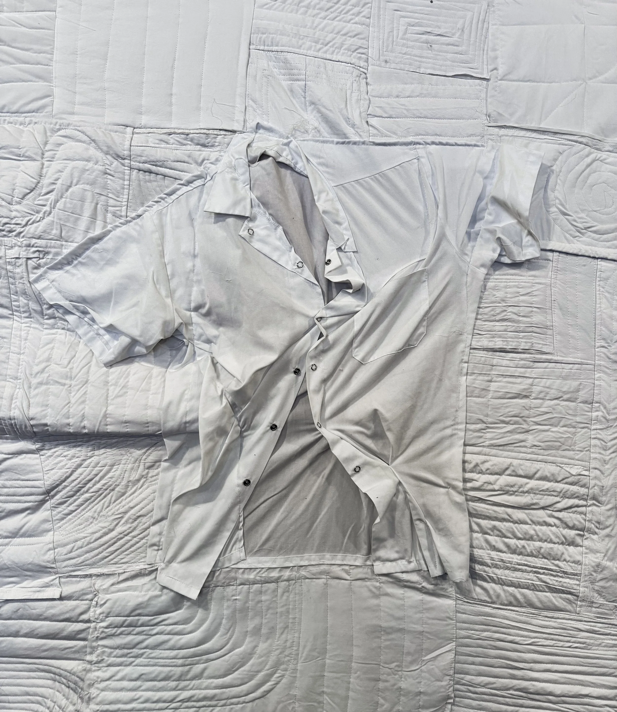 A white dress shirt with black buttons is spread out on a bed with a textured quilted bedspread.