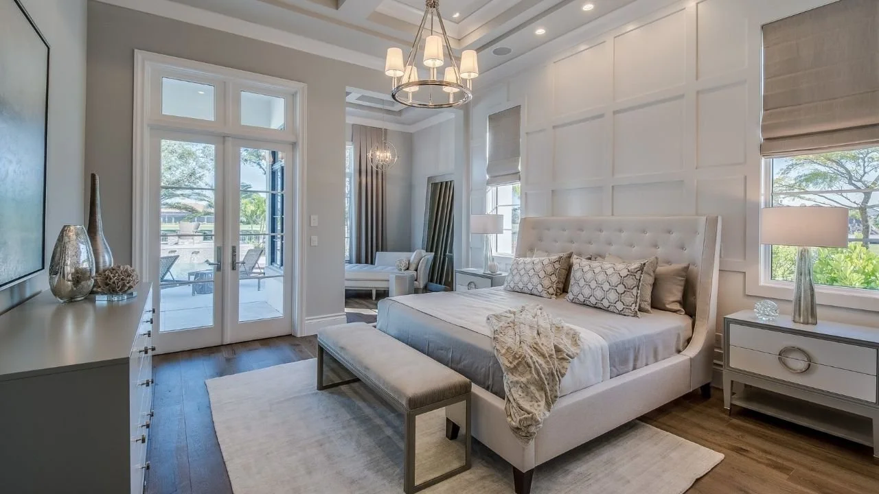Image of a master bedroom with a large white statement wall, chandelier, and fully made with extra pillows bed.