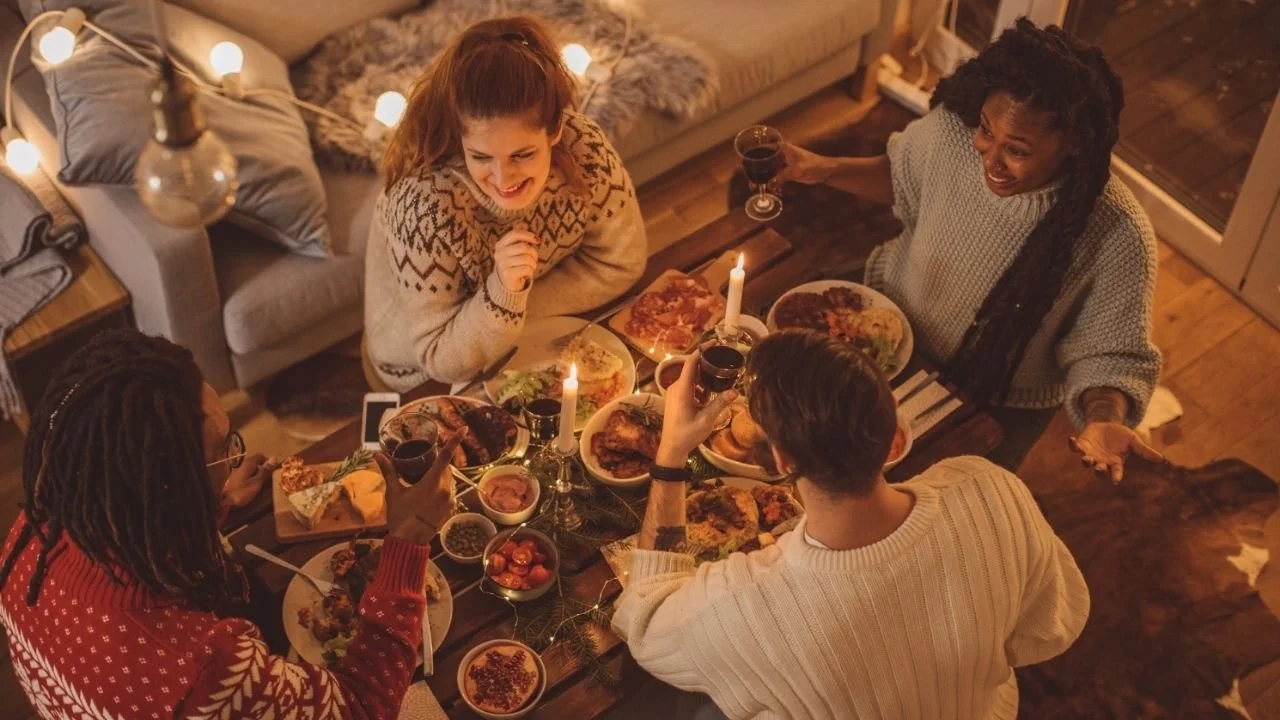 Image with view from above of a group of 4 people sitting around a small table full of food. They are all drinking wine and smiling and laughing.