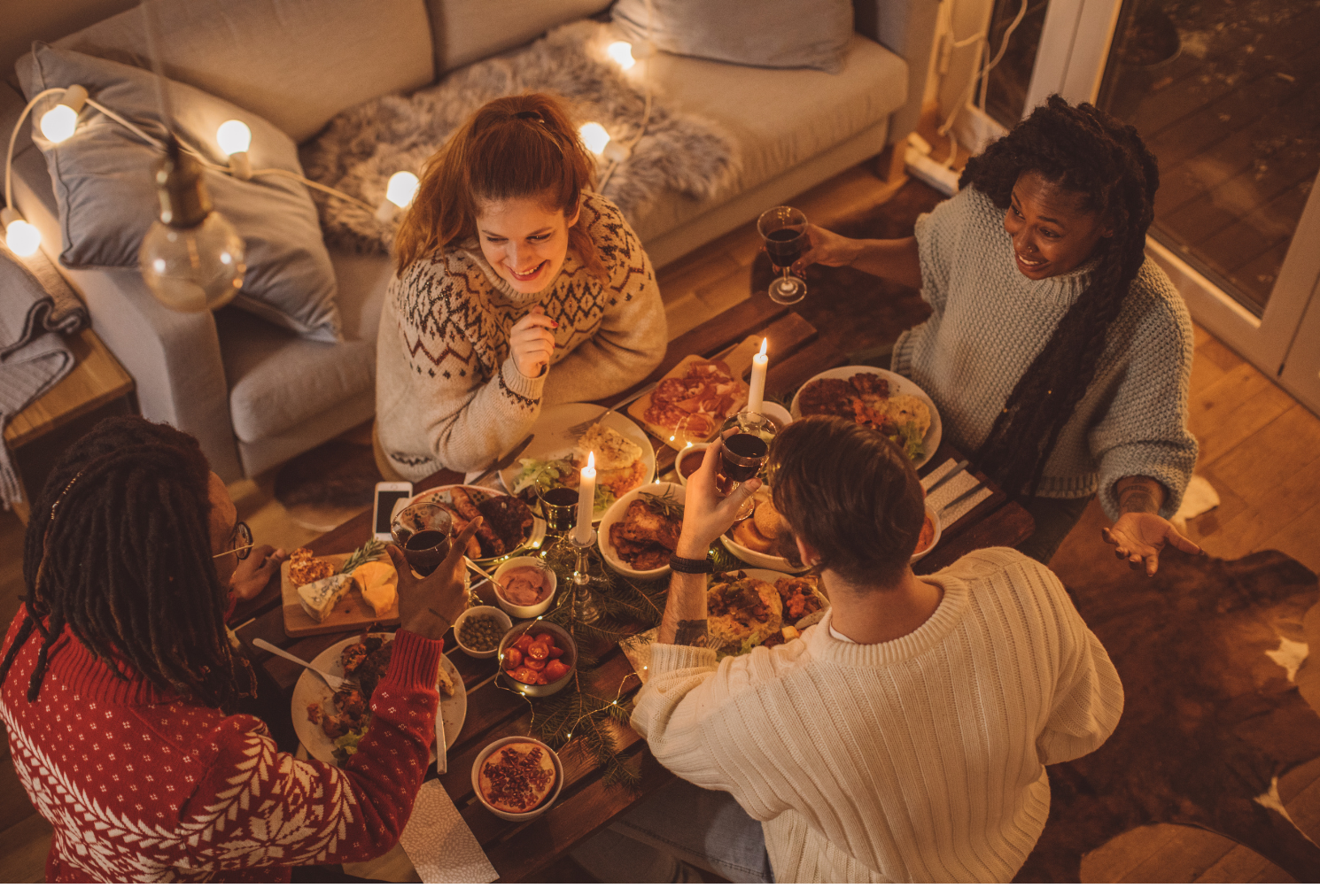Image with view from above of a group of 4 people sitting around a small table full of food. They are all drinking wine and smiling and laughing.