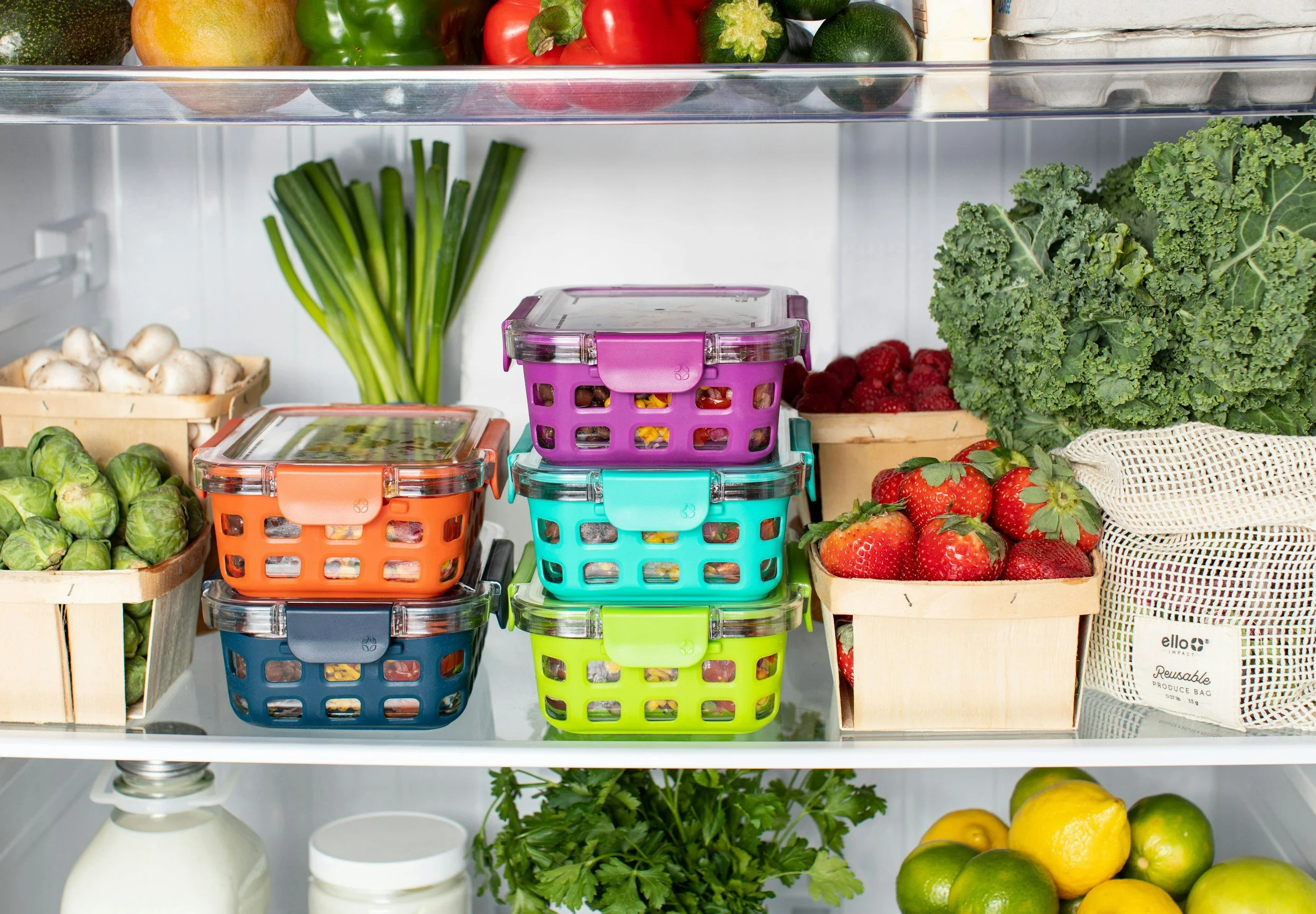 Inside a fridge sits colorful containers full of food and surrounded by fresh fruits and vegetables like strawberries and lemons.