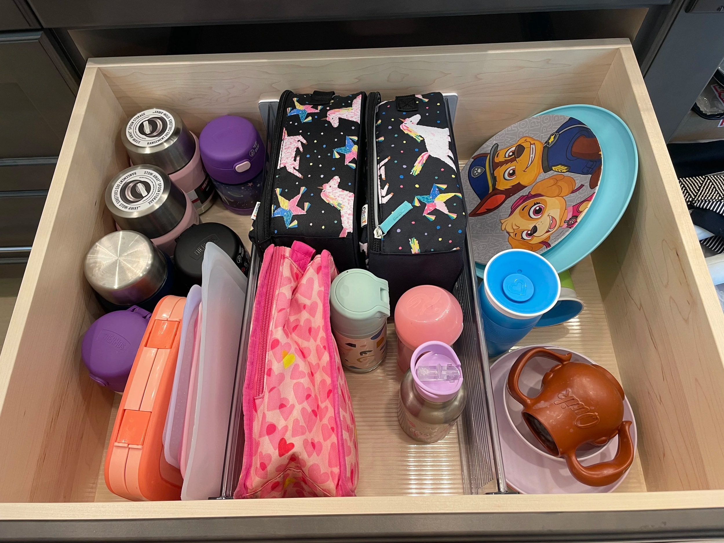 An open kitchen drawer with clear drawer dividers holding one section of lunch boxes, soup containers and bottles and another divider separating the regular kid dishware.