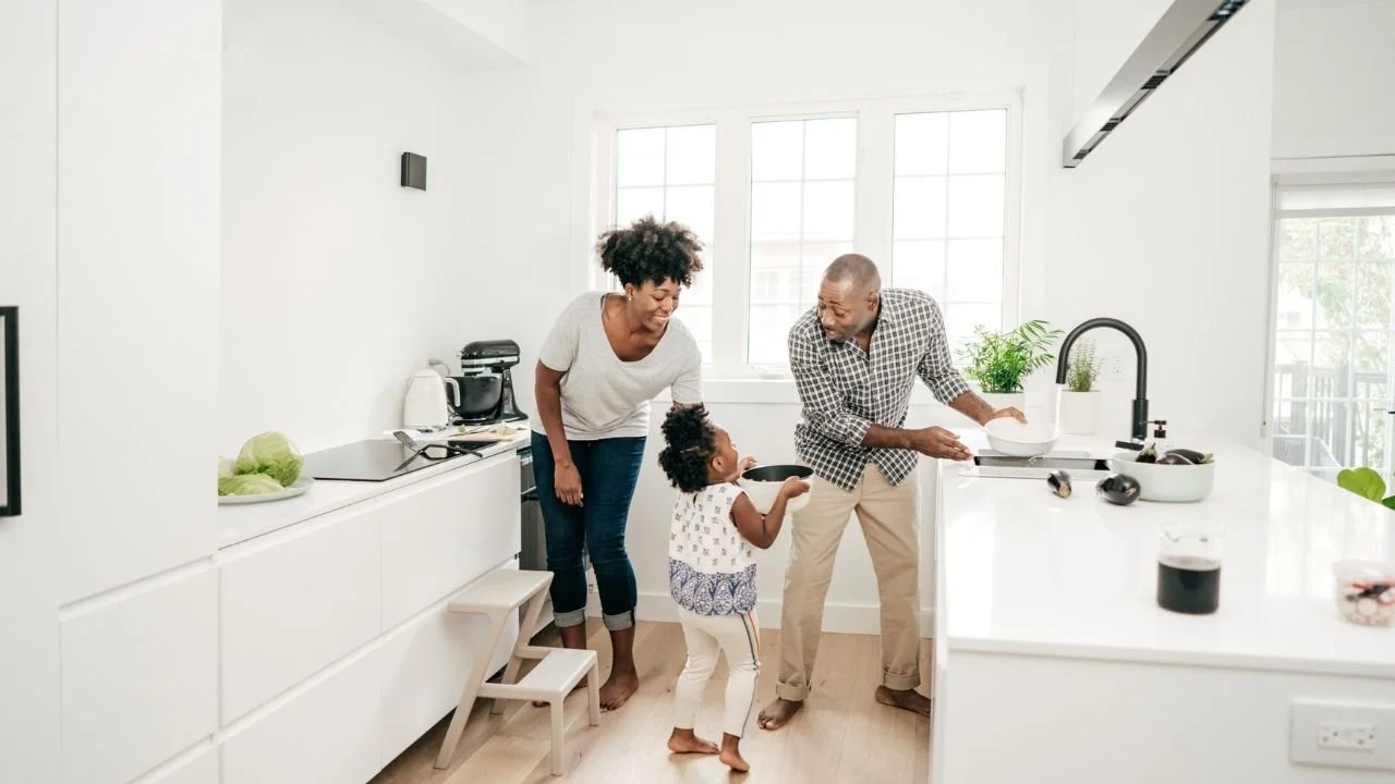 Image of a family  working together in an all white kitchen. They are passing dishes around to the sink and looking at each other happily.