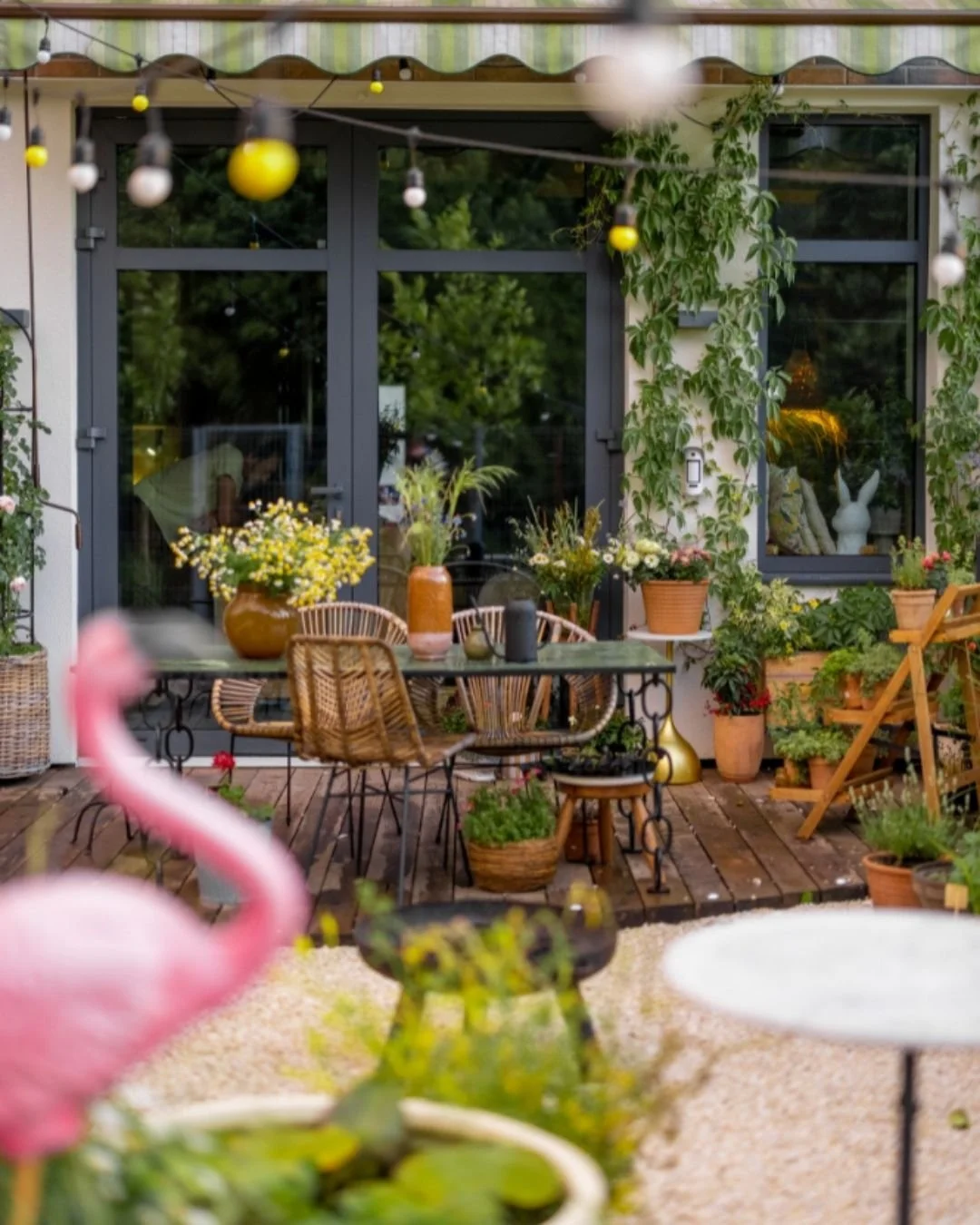 Backyard patio with table and chairs, many potted flowers and plants with both handing lights and a plastic flamingo in the foreground.