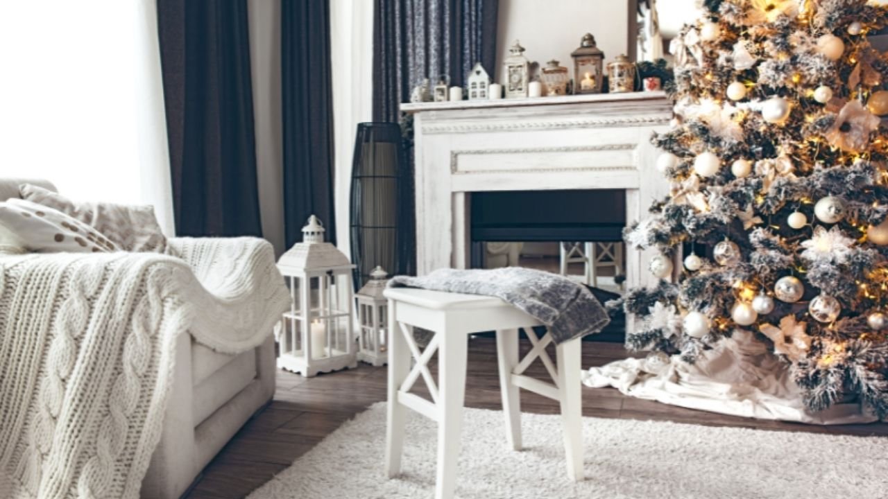Off-white sitting room with black sheer curtains, a big comfy chair with a white wool blanket near a fireplace and decorated Christmas tree with white, silver, and gold ornaments.