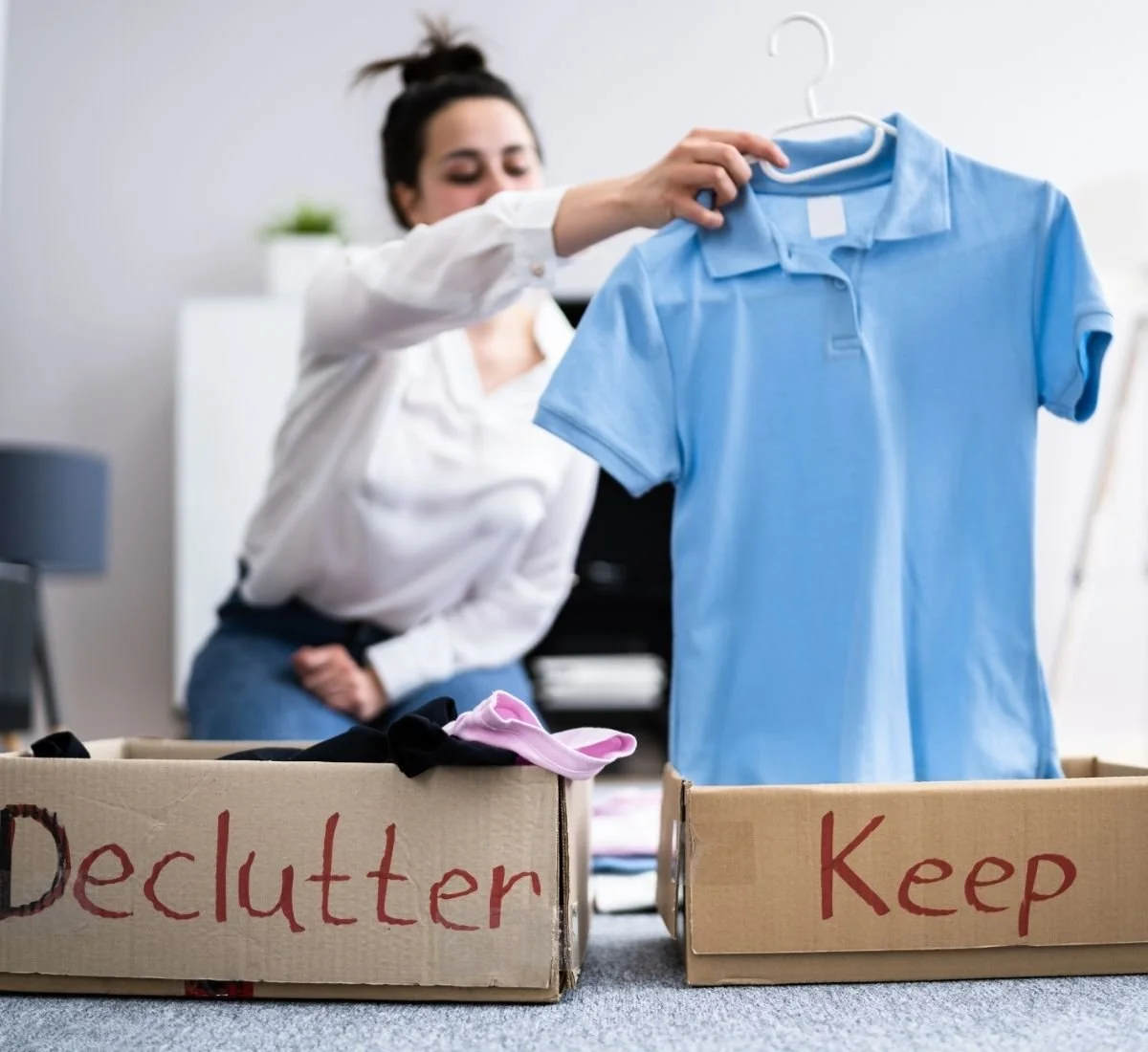A woman with dark hair in a bun wearing a white button up shirt and blue jeans, holding a hanger with a men's light blue collared polo. She is putting it in a box labeled "Keep" not the other boxed labeled "Declutter"