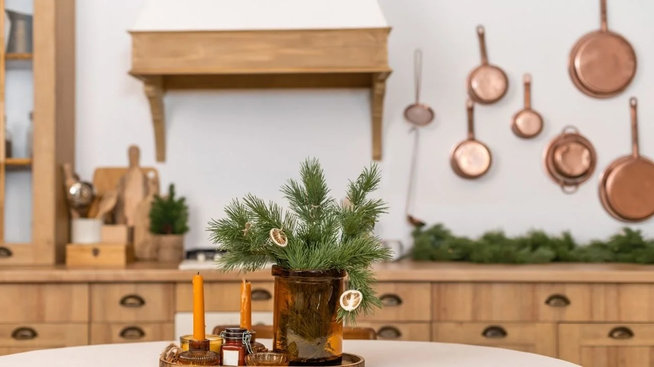 Image of a brown wood kitchen. On the island is a red vase filled with evergreen branches and dried oranges. Surrounding the vase are multiple orange candle sticks