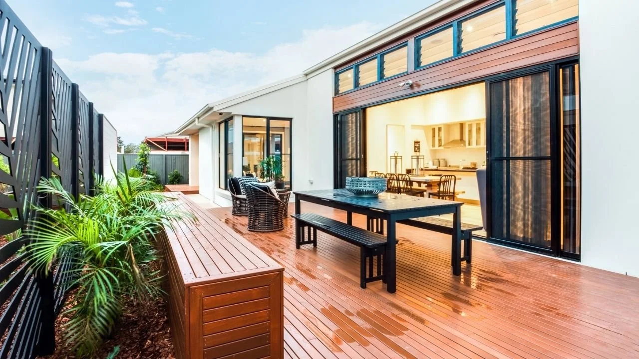 Image of a wood composite deck with a dining table and sitting area, surrounded by a black fence, plants, and open to the inside kitchen