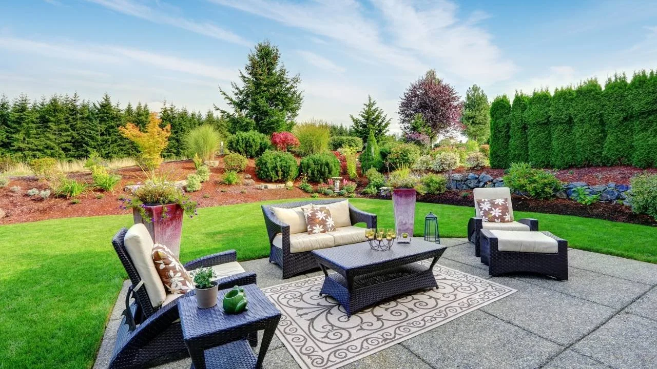 Backyard patio with patio furniture and a bright green lawn surrounded by tall trees, full bushes, and grasses.