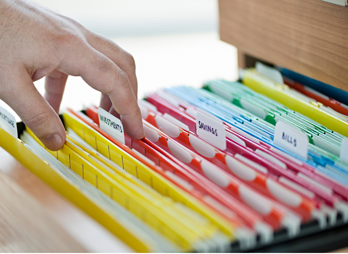 Image of a white hand sorting through an open file drawer filled with file folders organized by color: yellow, orange, red, blue, and green. The folders are full of paperwork and the files are labeled with tabs labeled "investments" and "savings".