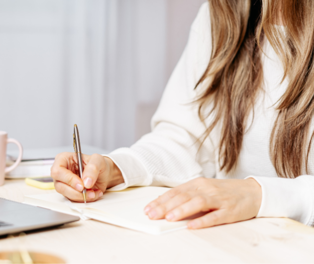 Person writing in a notebook at a desk with a laptop and mug nearby