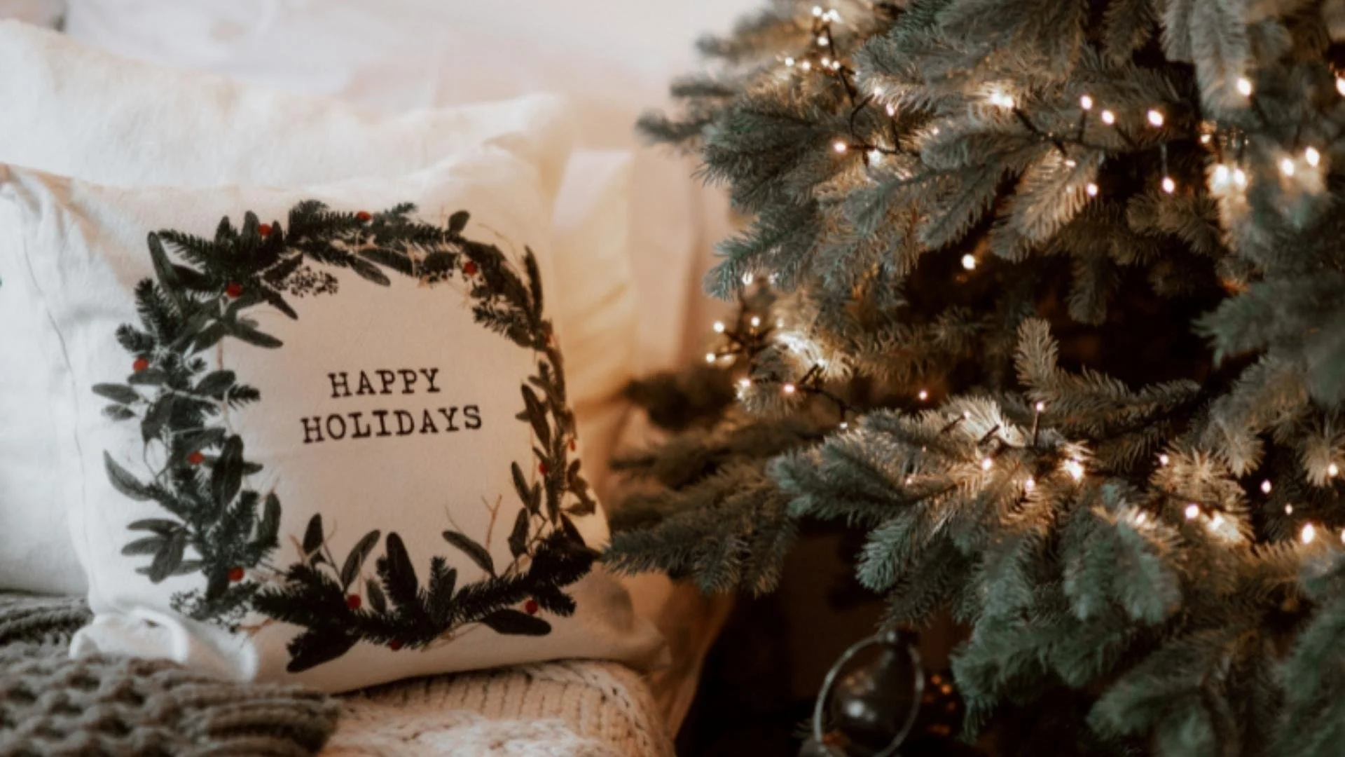 Image of a bed with many pillows, a Happy Holidays encased in a wreath print pillow, a green wool blanket and to the right is a simple Christmas decorated with white twinkle lights and minimal ornaments