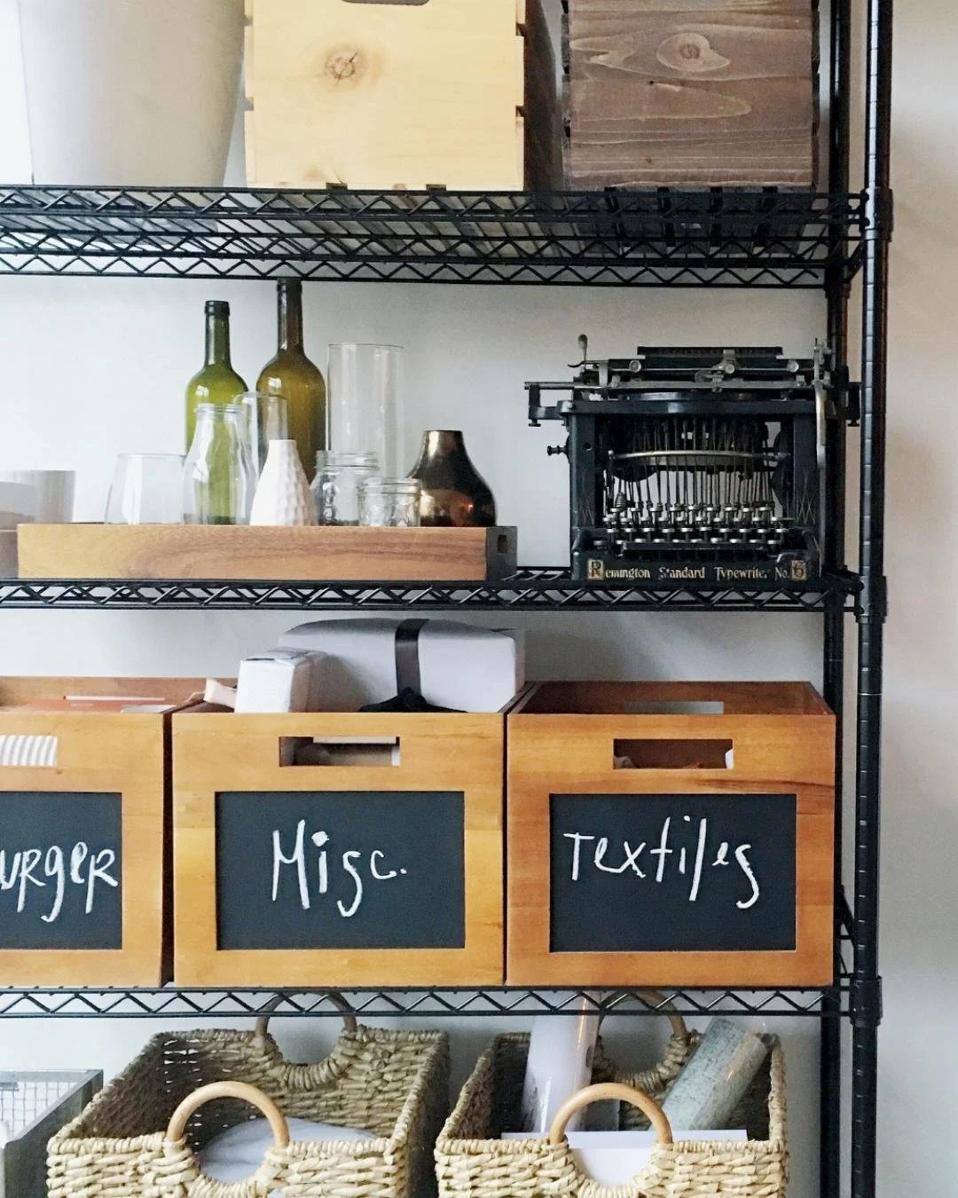 Image of a black metal rack holding different baskets and trays. The tray holds empty vases and under that are wooden baskets with labels written in chalk like "Misc" and "Textiles"